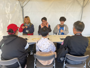 本学産業技術学部の学生3名が、ブースを訪れた一般の方3名と向かい合って座り、それぞれ手話を教えている様子