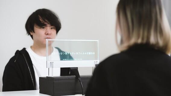 透明ディスプレイを挟んで向かい合う2人の人物。手前の人物は後ろ姿で、奥の人物は正面を向いている。2人の間に置かれた透明なアクリル板状のディスプレイに日本語のテキストが両面に表示されており、会話の字幕として機能していることが伝わる。