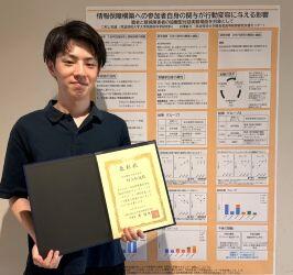 ポスター発表内容の前で表彰状を持っている村上さんの写真